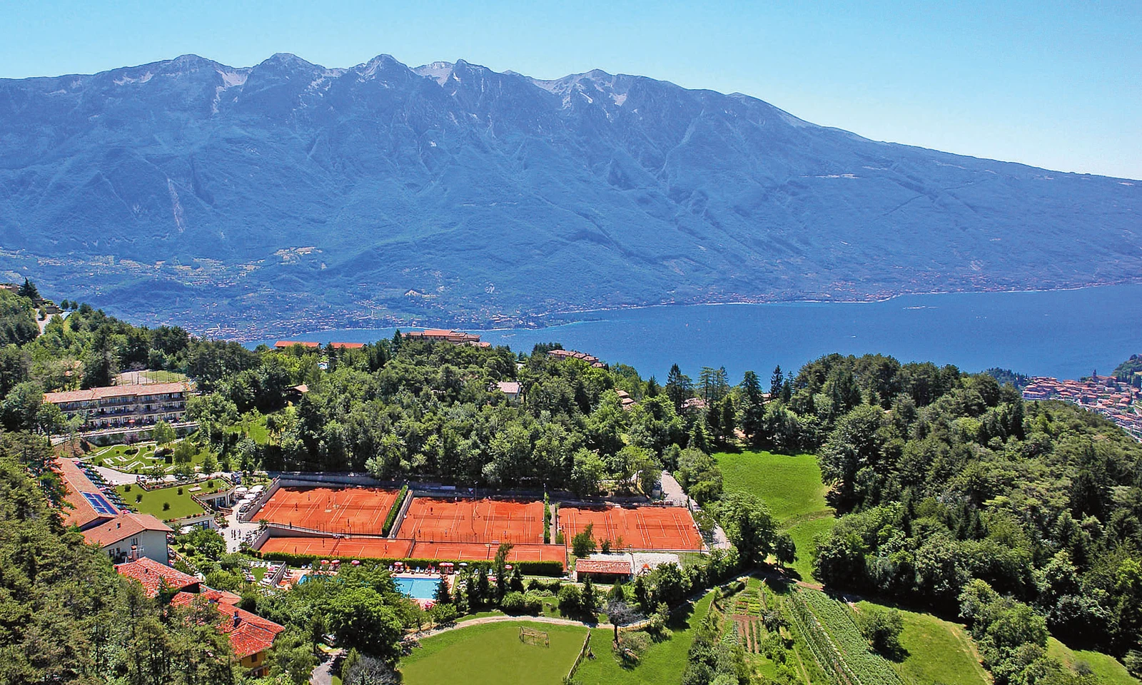 Tennis courts, surrounded by lush greenery and buildings, with a lake and mountains in the background at Residence Campi