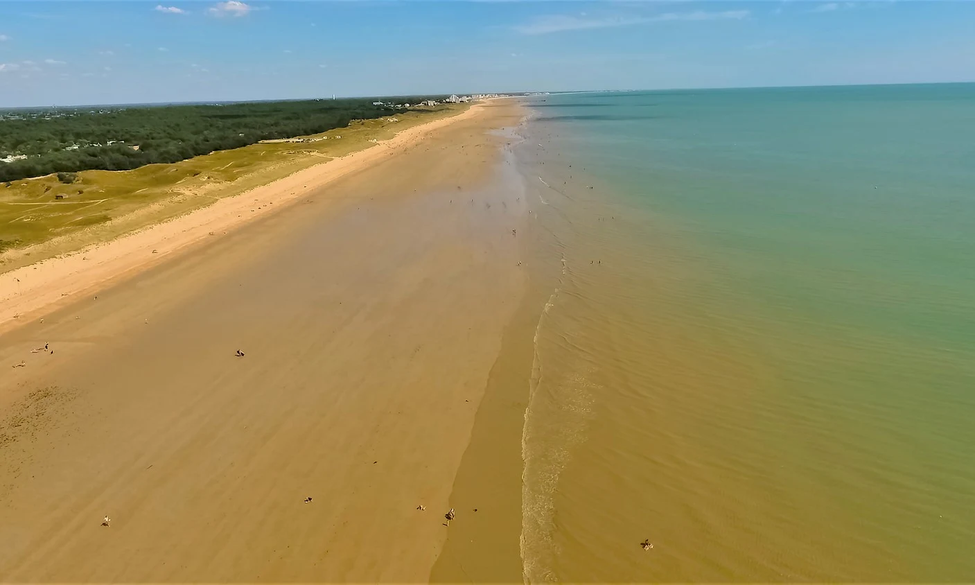 Expansive beach with people scattered, walking along the water's edge; grassy dunes and forest line the background at Les Aventuriers de la Calypso