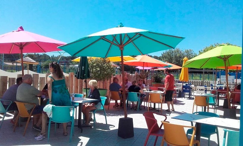 People sitting and chatting under colorful umbrellas with scattered tables and chairs on a sunny patio at Les Aventuriers de la Calypso