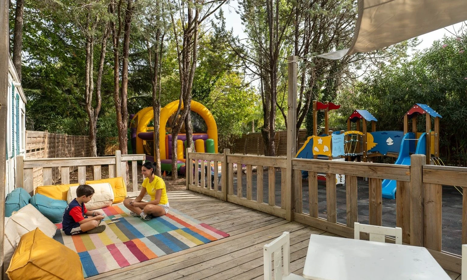 Two people sit and play on a colorful rug on a wooden deck near a playground, surrounded by trees at La Barque