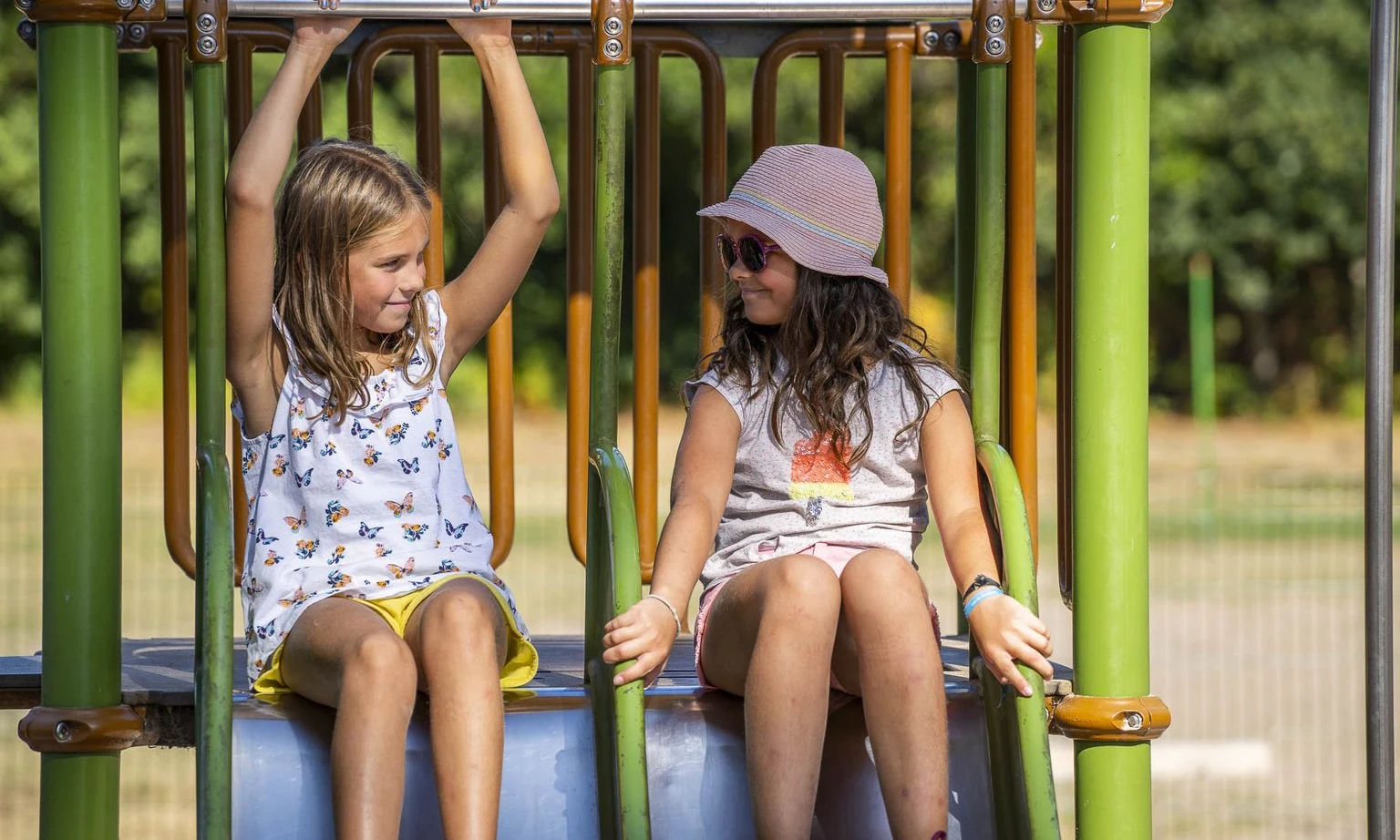 Two girls sitting on playground equipment, one hanging from a bar, outdoors at Lac de Sanguinet