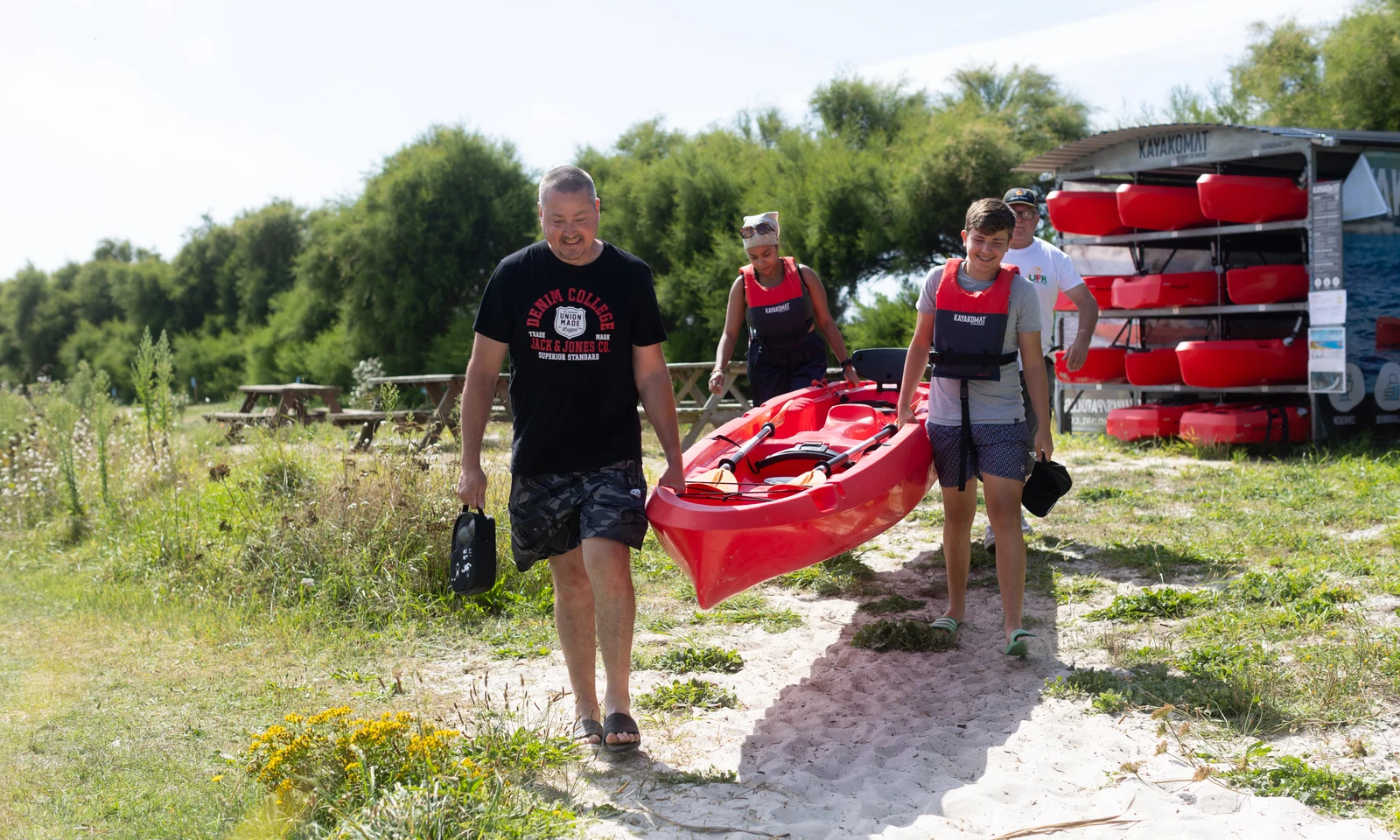 People carrying a red kayak, walking on a sandy path beside stacked kayaks and picnic tables, surrounded by greenery at La Pointe de Roscoff