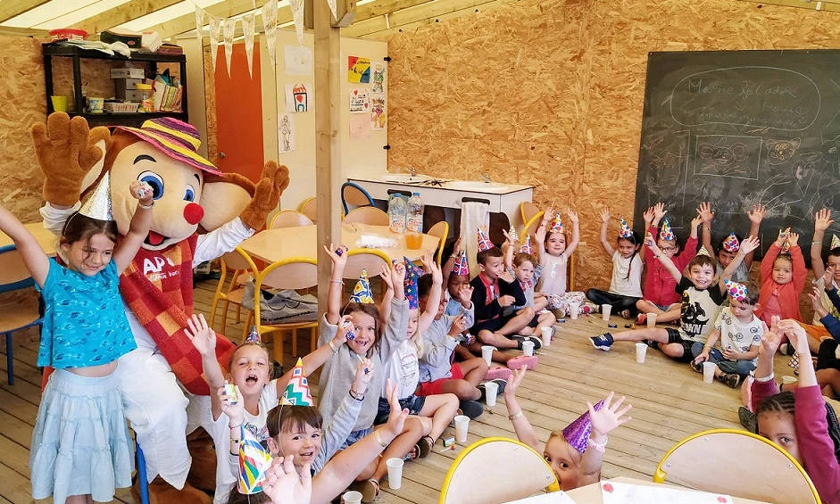 Children wearing party hats raise hands, seated around mascot in colorful, decorated indoor space at Les Aventuriers de la Calypso