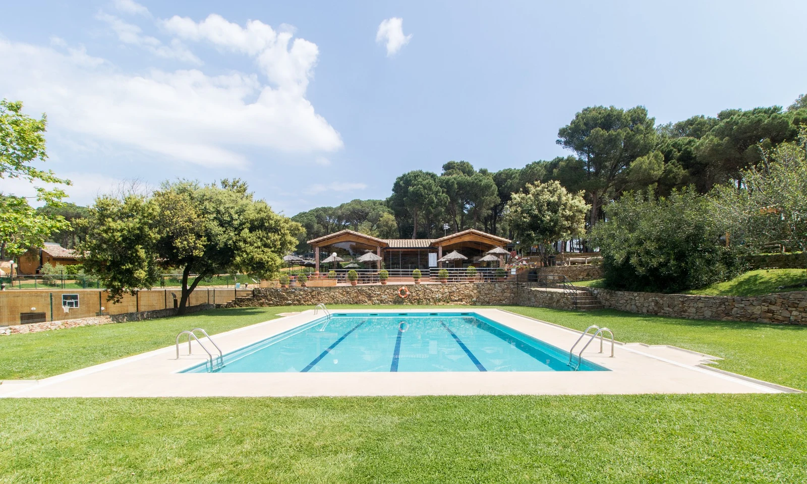 Swimming pool with ladders, surrounded by grass and trees, with buildings and seating areas in the background at Begur