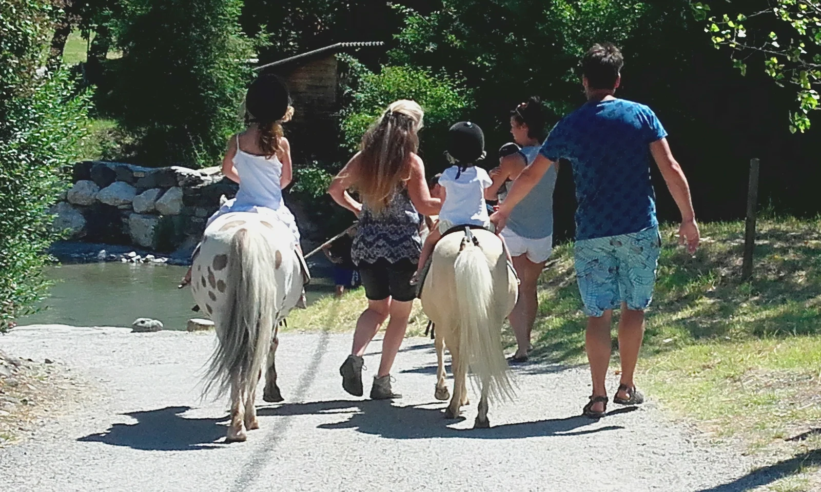 Children riding ponies, accompanied by adults, on a wooded path beside a river at Les Arches