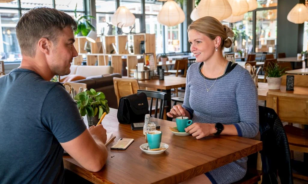 Two people converse at a wooden table, sitting with coffee cups in a well-lit café with modern decor at Landal Rabbit Hill