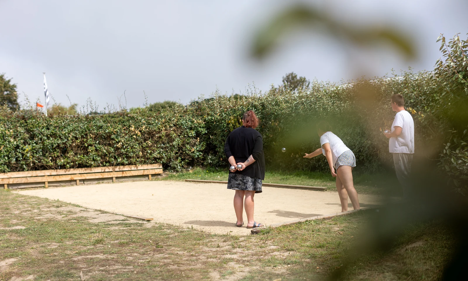 People playing pétanque on a sandy court, surrounded by green hedges. One person throws a ball at La Pointe de Roscoff