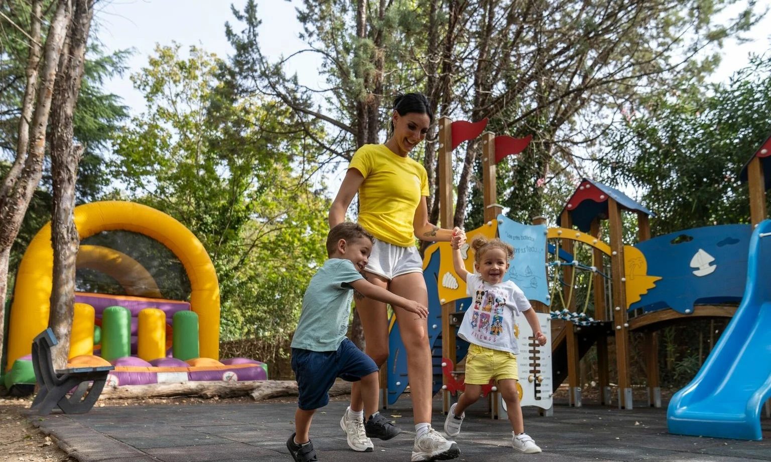 Woman holding hands with two jumping children on a playground, surrounded by trees at La Barque