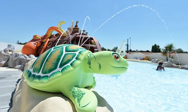 Green turtle statue with water jets in a children's pool area, with a large orange slide in the background at Les Aventuriers de la Calypso