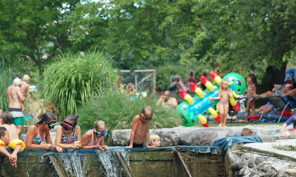 Children observe water flowing over wooden barriers, surrounded by trees and greenery at Les Arches