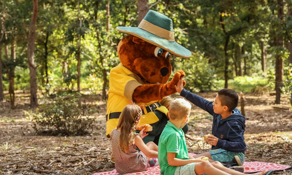 A person in a bear costume high-fives a child; other children sit on a picnic blanket among trees at Landal Rabbit Hill