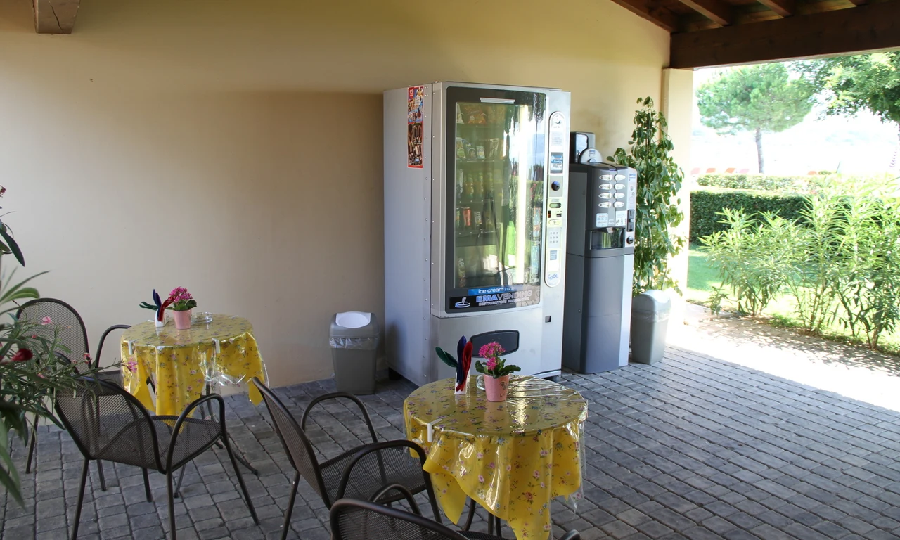 Vending machines beside a small seating area with yellow tablecloths and flowers, situated outdoors under a sheltered patio at Front Lake Resort Le Corti del Lago