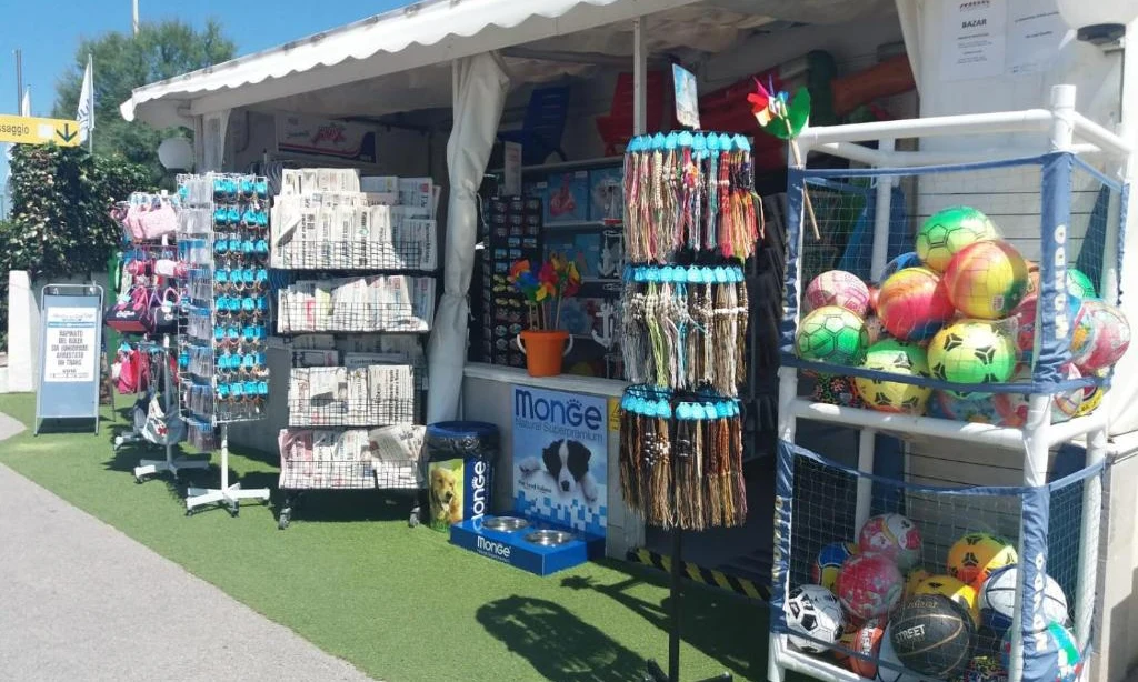 Outdoor stand displaying colorful balls, bracelets, and beach items on shelves; set on artificial grass at International Riccione Family Camping Village