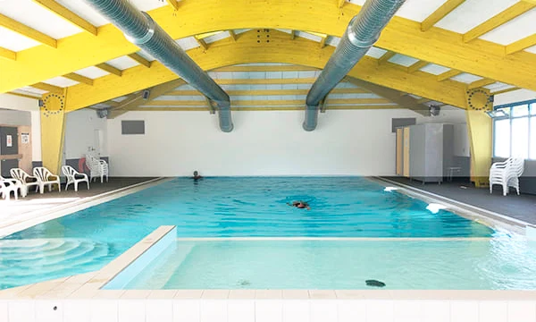 Indoor pool with a swimmer in a bright, spacious room with yellow ceiling beams and stacked chairs at Les Aventuriers de la Calypso