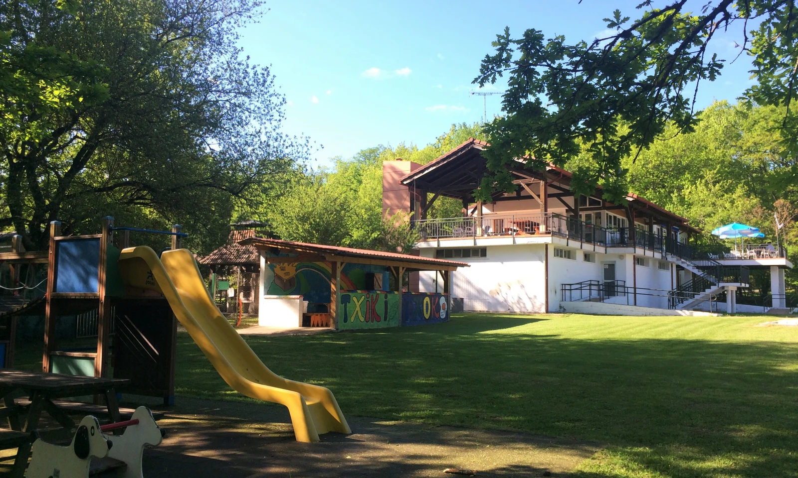 Playground slide sits empty amidst grassy area, near a building surrounded by trees under a clear sky at Etxarri