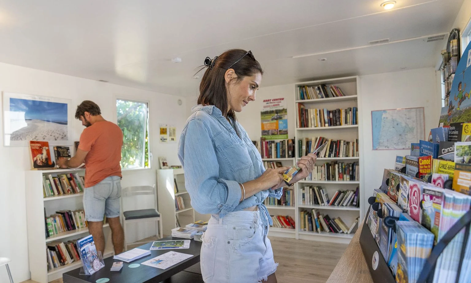 Two people browse books in a brightly lit, cozy library with shelves and brochures at Lac de Sanguinet