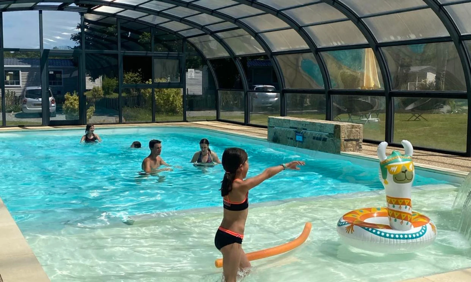 A child plays with a pool noodle in a covered swimming pool, with others enjoying the water around at Domaine de Mesqueau