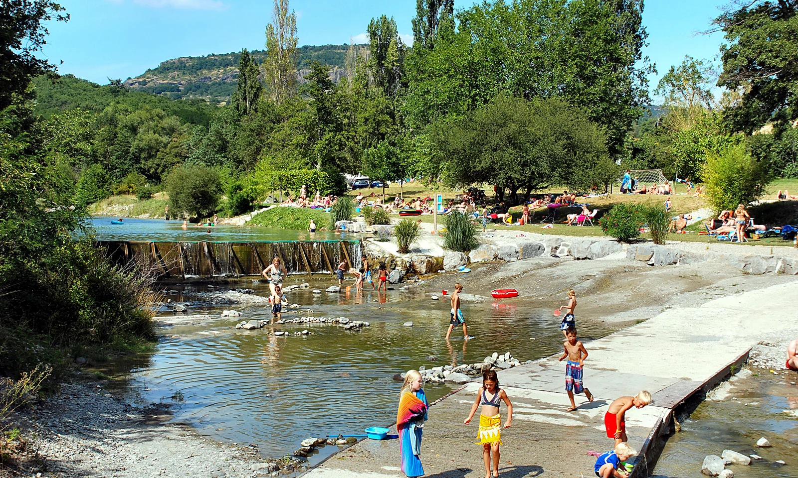 Children play in a shallow river while people relax on grassy banks, surrounded by lush trees at Les Arches