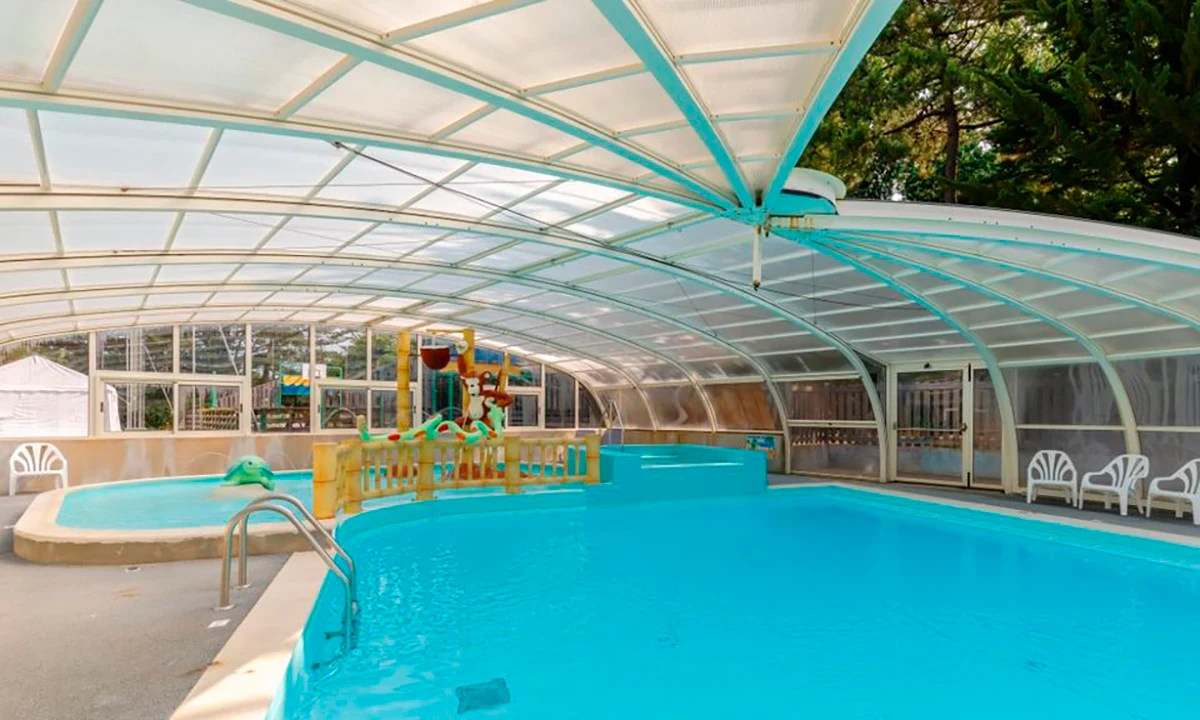 Covered swimming pool with slides and a play structure, surrounded by chairs at Camping 2 Plages & Océan Village Corsaire