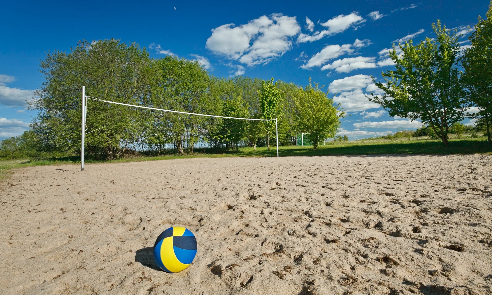 A volleyball rests on a sandy court with a net, surrounded by trees and under a partly cloudy sky at KNAUS Campingpark Hünfeld