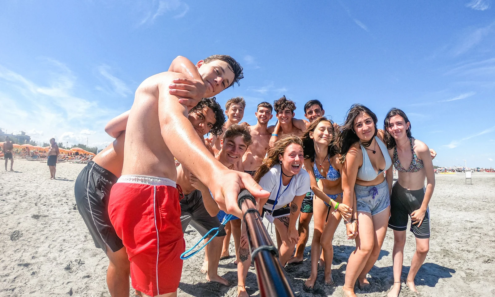 A group of smiling young people poses with a selfie stick on a sunny beach, surrounded by sand and umbrellas at International Riccione Family Camping Village