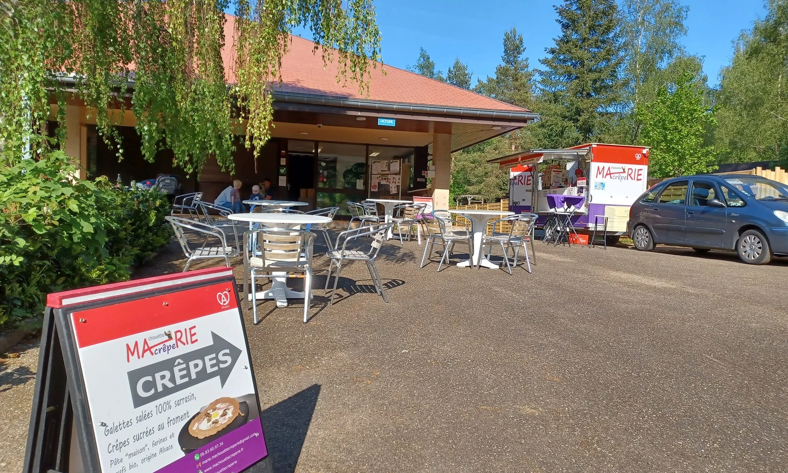 Outdoor tables with chairs sit empty beside a crepe food truck, surrounded by trees and a building at La Foret