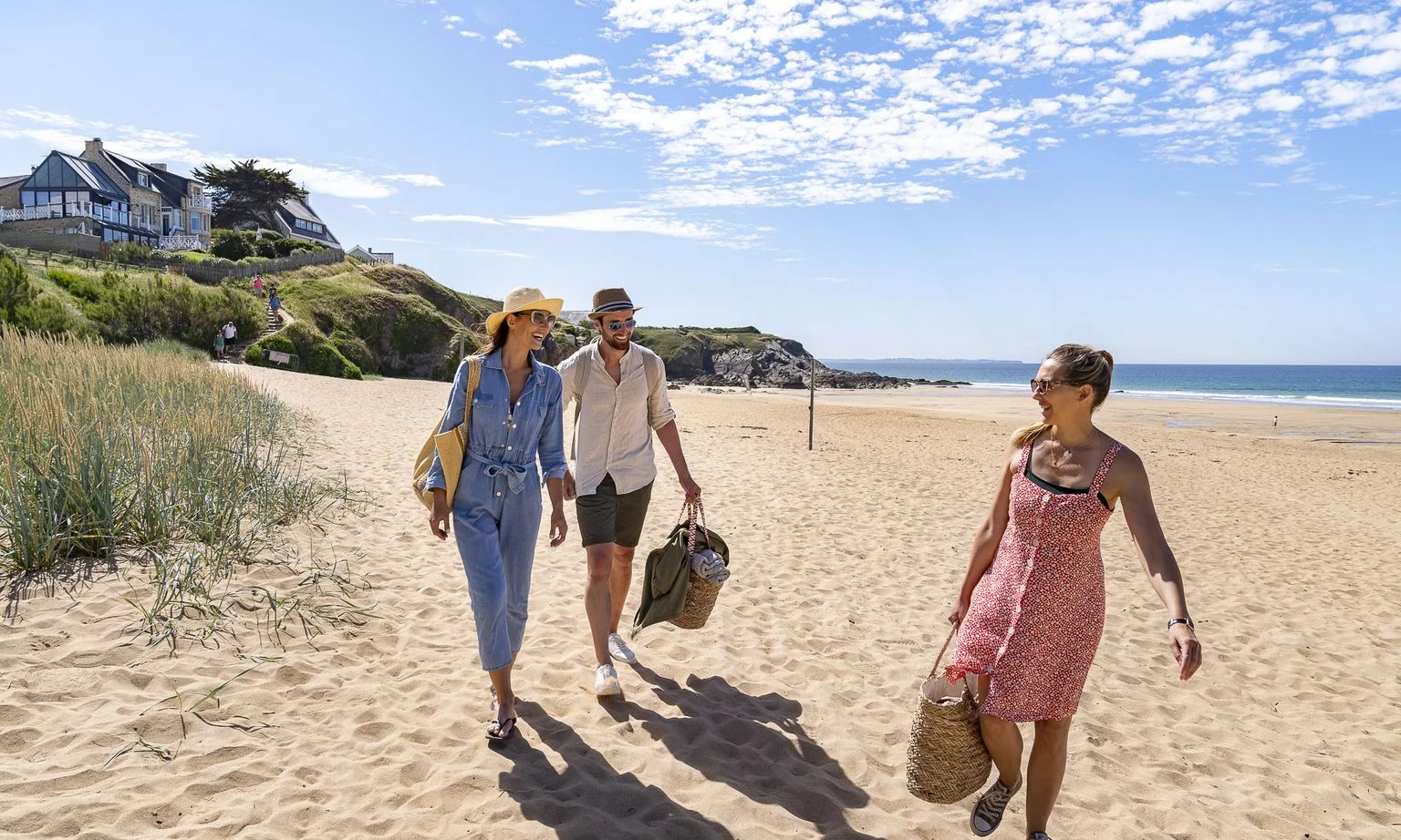 Three people walk on a sandy beach carrying bags near a grassy hill with houses, under a bright blue sky at Lac de Sanguinet