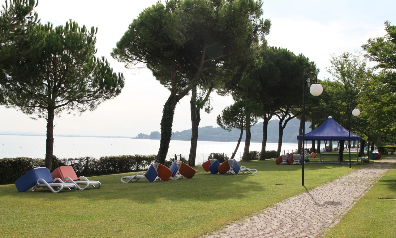 Sun loungers arranged under trees beside a lakeside pathway, with a purple canopy in the background at Front Lake Resort Le Corti del Lago