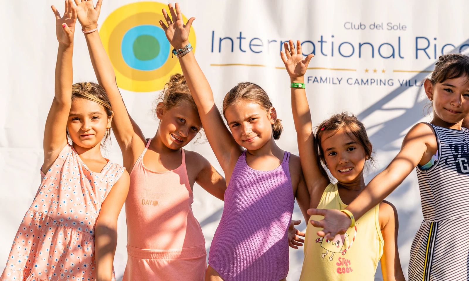 Four children smiling, raising arms, in bright outfits against a sunny backdrop, enjoying outdoor activities; at International Riccione Family Camping Village