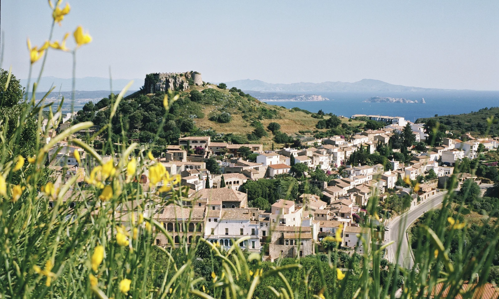 Ruins of a stone fortress on a hill overlook a village near a coastline, surrounded by yellow wildflowers at Begur