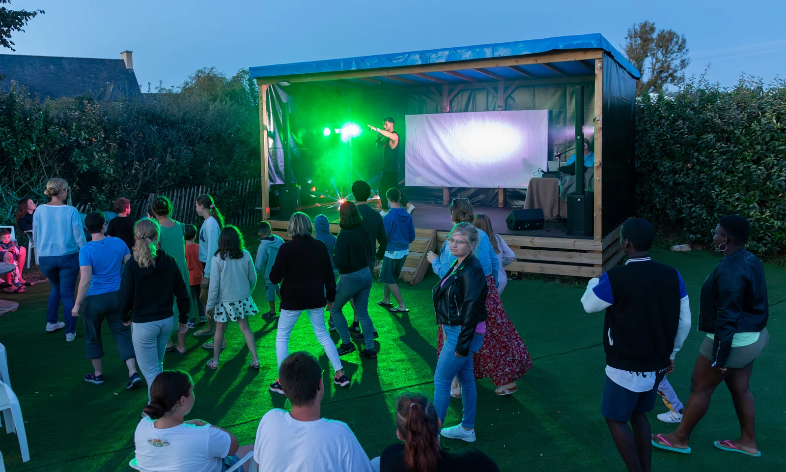 Performer sings on a stage with green lights, entertaining a group of people gathered outdoors at La Pointe de Roscoff