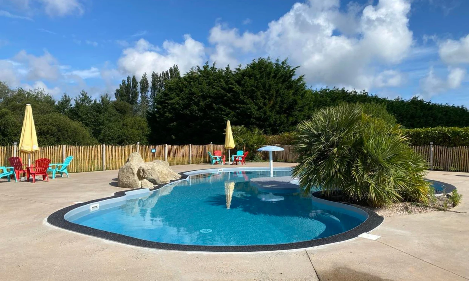 A small pool surrounded by lounge chairs and umbrellas, with lush greenery and a vibrant blue sky at Domaine de Mesqueau