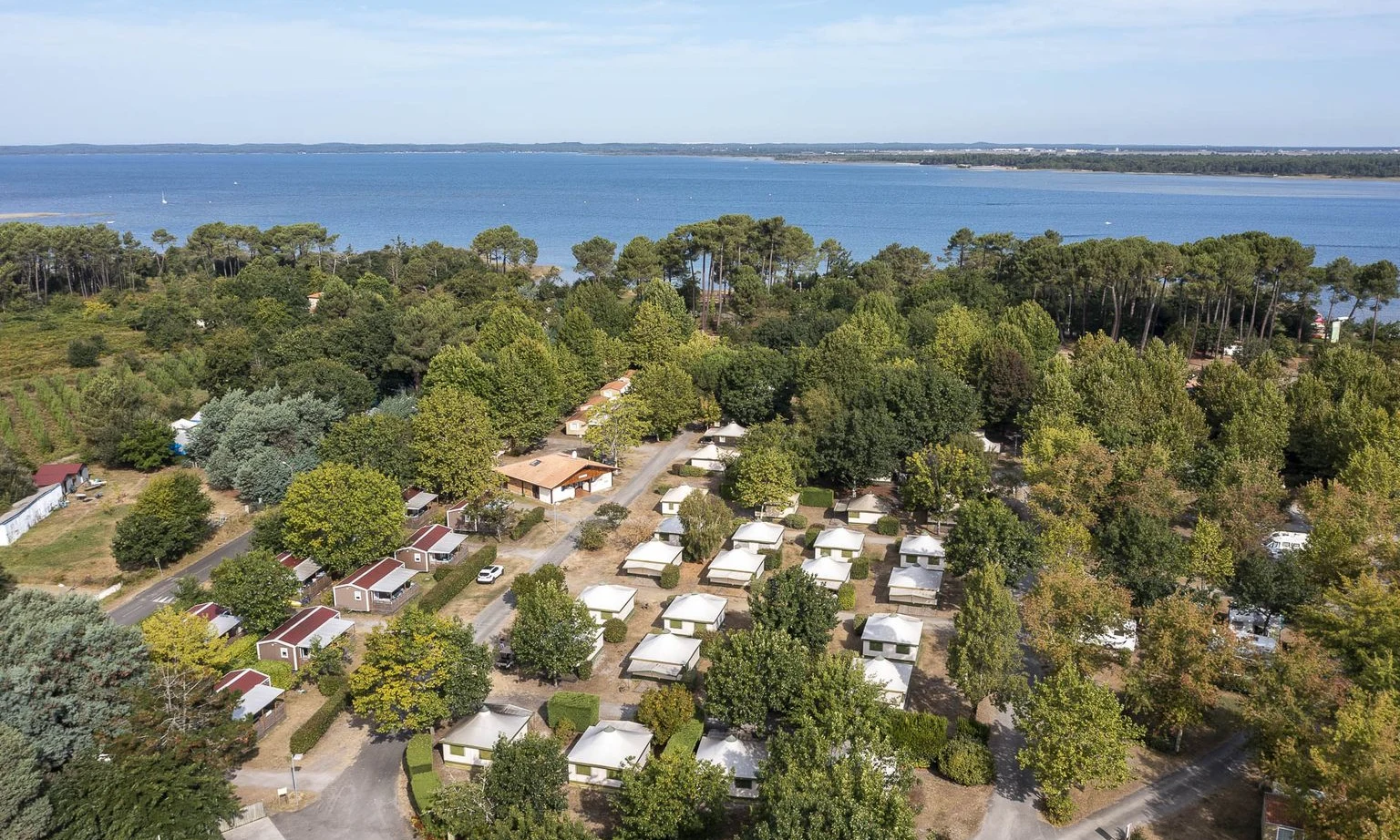 Cabins and tents nestled among trees beside a broad lake, with roads connecting various sections at Lac de Sanguinet