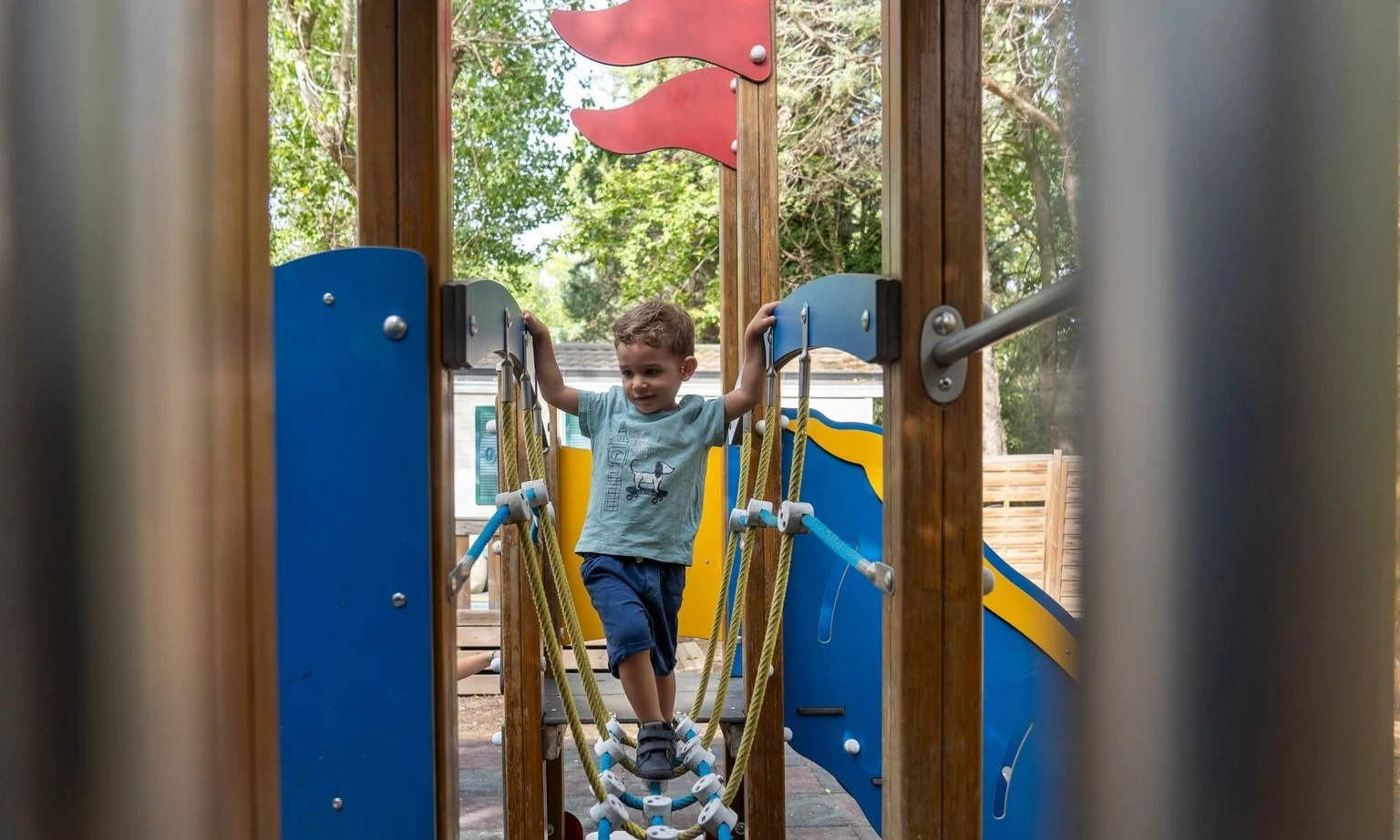 Child crossing a rope bridge on a playground, surrounded by trees and structures at La Barque