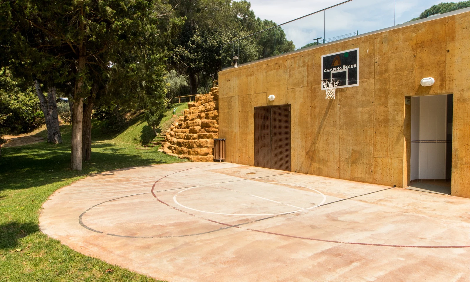 Basketball hoop mounted on a concrete wall beside a half-court basketball area, surrounded by trees and rock steps at Begur