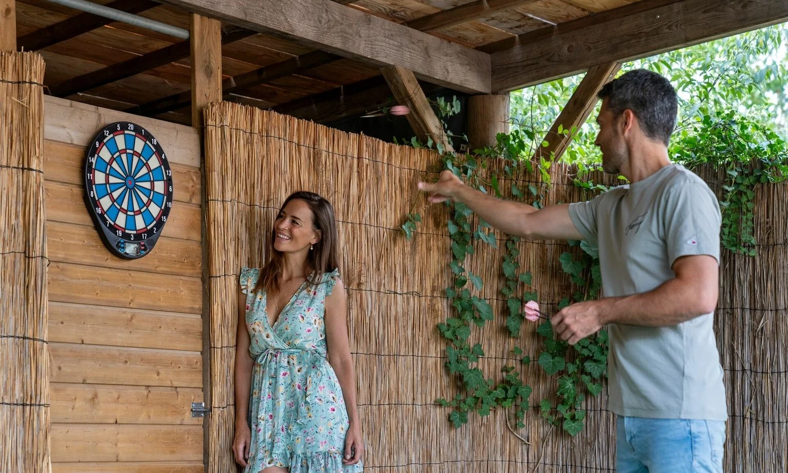 Man throws darts, woman watches, standing beside an outdoor dartboard, in a wooden thatched-roof structure with greenery at La Barque