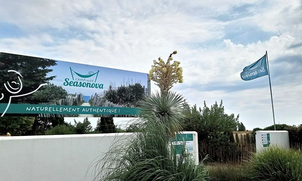 A sign and flag display a campsite’s logo and slogan, surrounded by greenery and beneath a cloudy sky at Seasonova Ile de Ré