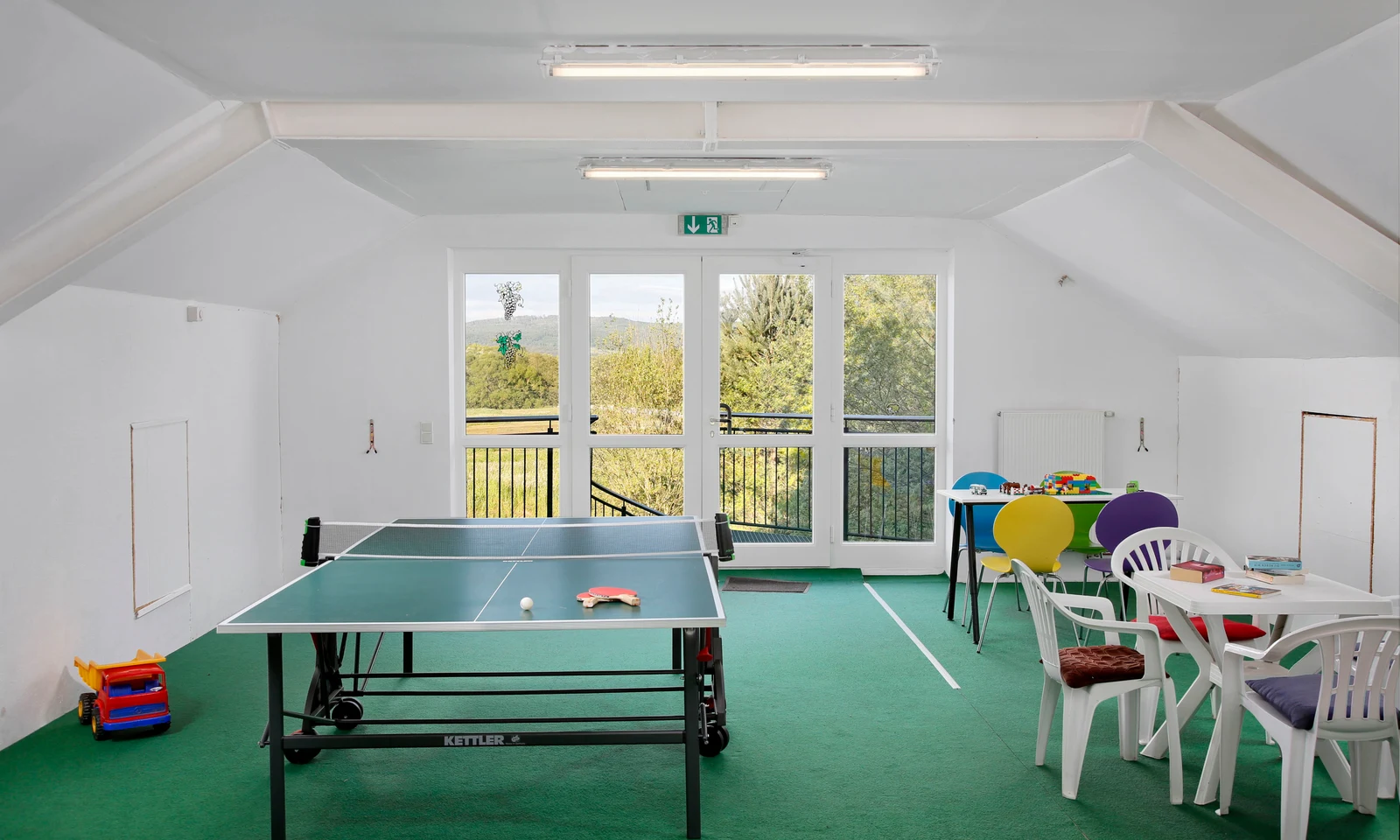 Ping pong table, chairs, and toys in a bright room with windows showing outdoor greenery at KNAUS Campingpark Hünfeld