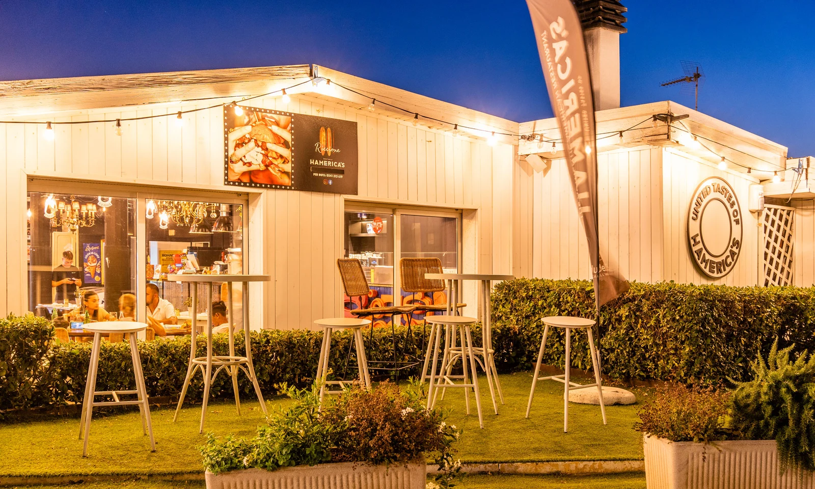 Outdoor tables and chairs arranged on green grass, overlooking a lit-up restaurant facade at International Riccione Family Camping Village