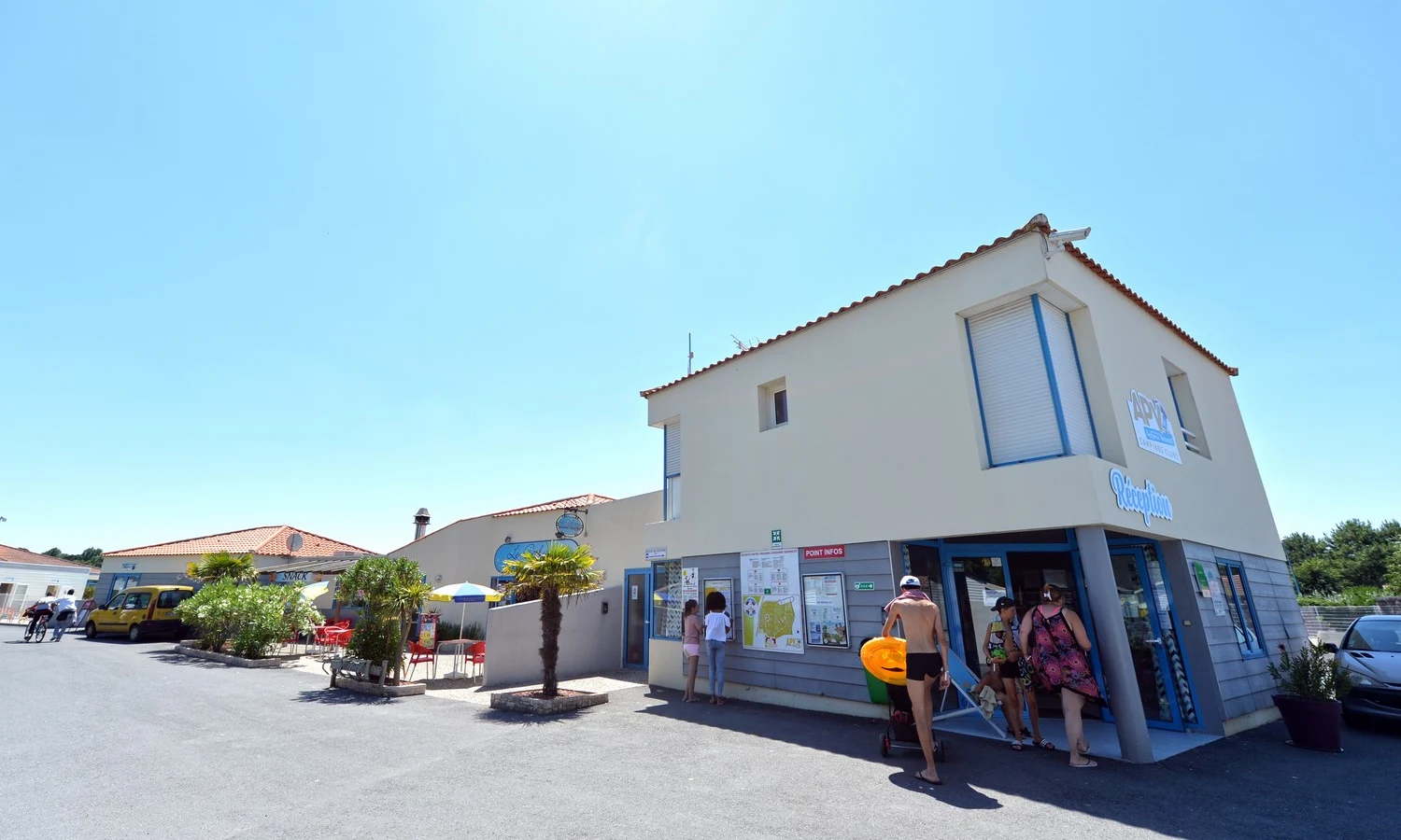 People entering a reception building; another person reading signs; outdoor tables nearby; blue sky background at Les Aventuriers de la Calypso