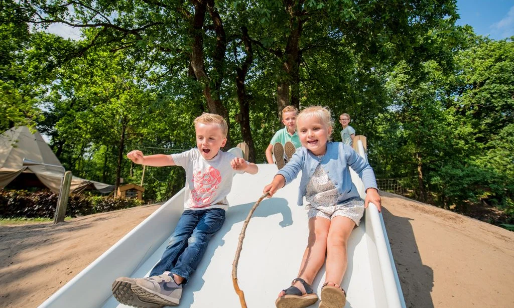 Children sliding down a playground slide, surrounded by trees and tents in a forested campsite setting at Landal Rabbit Hill