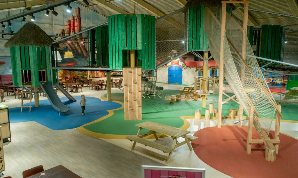 Indoor playground with slides, climbing nets, and elevated green huts, surrounding seated areas and child walking at Landal Rabbit Hill