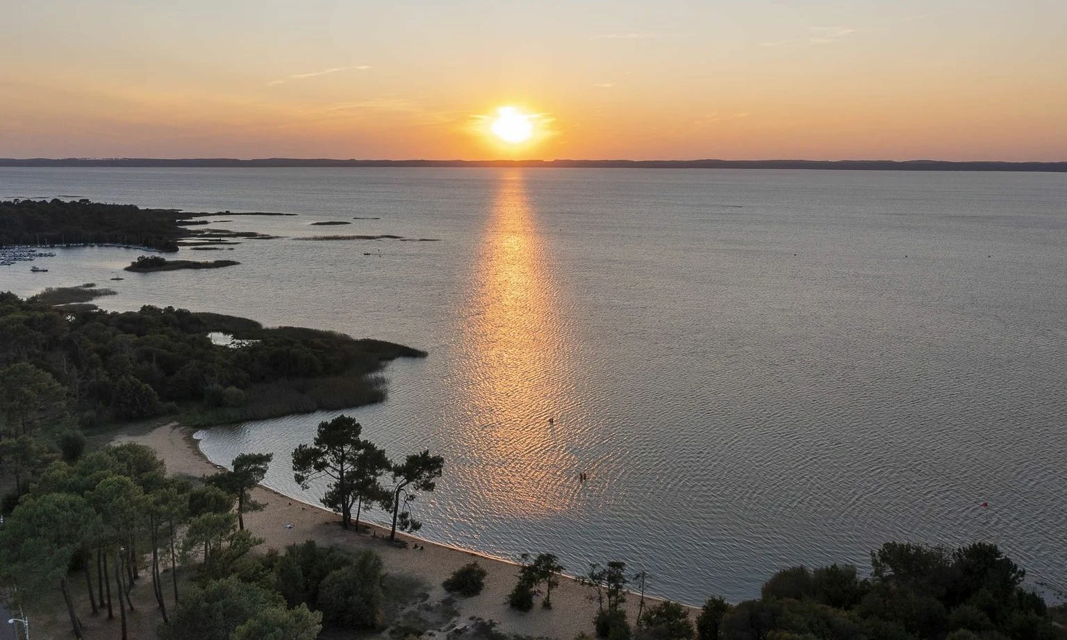 Sun setting over a vast lake, with a sandy shoreline bordered by dense trees at Lac de Sanguinet