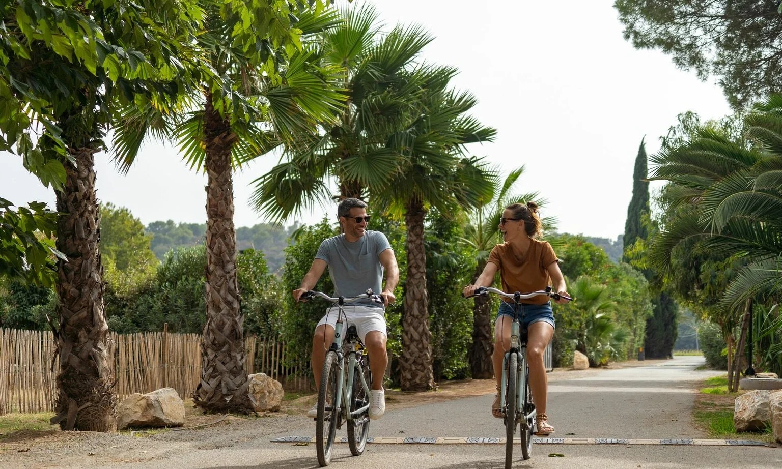 Two people cycling along a pathway lined with palm trees and greenery, smiling and enjoying the outdoors at La Barque