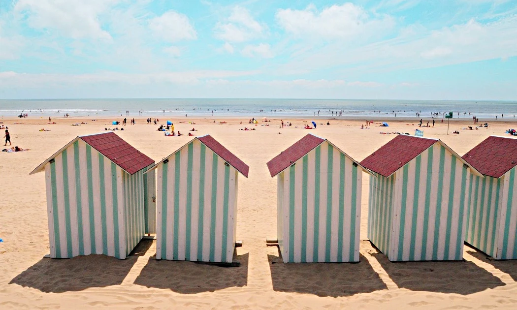 Striped beach huts lined up, facing the ocean, amongst scattered beachgoers enjoying the sandy shoreline at Les Aventuriers de la Calypso