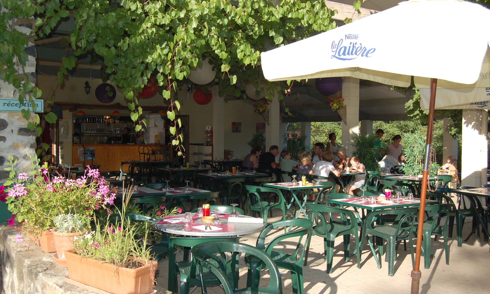 Patio seating area with green chairs under leafy canopy, people dining near a bar counter at Les Arches