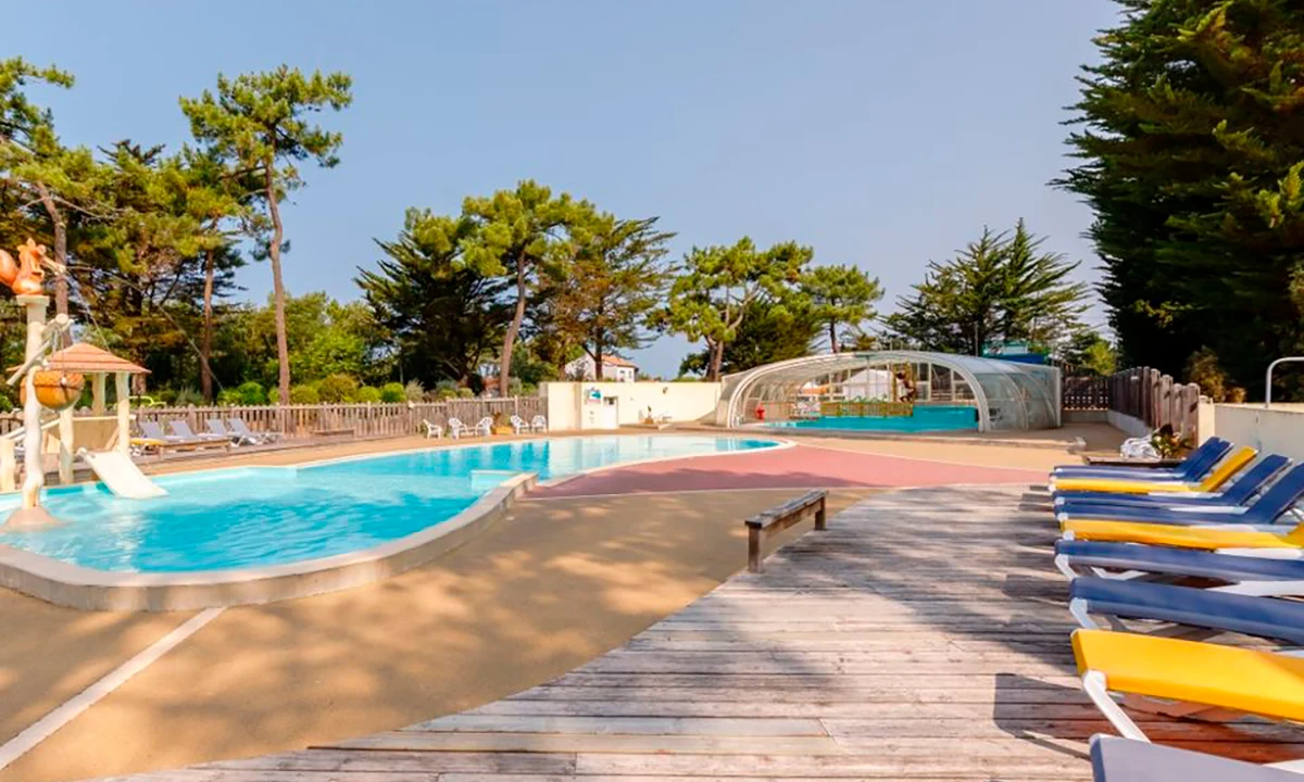 The swimming pool, with lounge chairs on a deck, is situated amongst trees and bordered by a covered pool area at Camping 2 Plages & Océan Village Corsaire