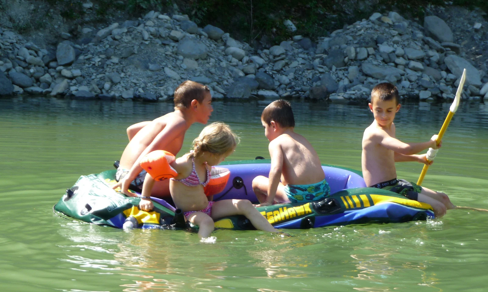 Children sitting on an inflatable raft paddle in a rocky river, surrounded by lush greenery at Les Arches