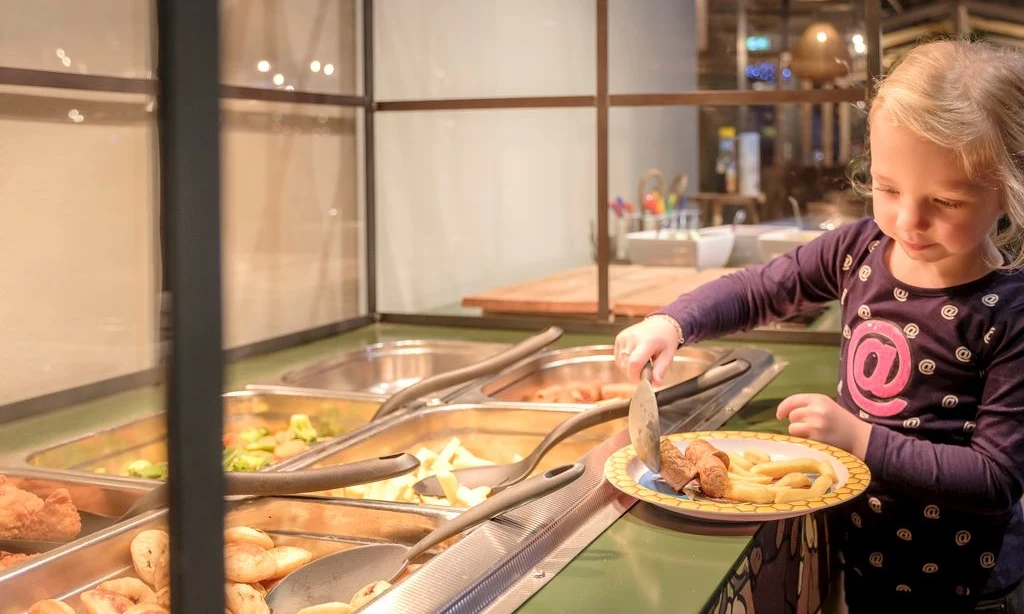 Child serving food at a buffet-style restaurant, holding a plate in a brightly lit indoor setting at Landal Rabbit Hill