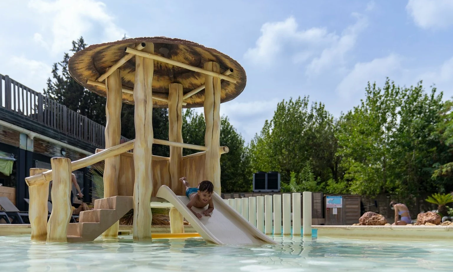 Child sliding down wooden playground slide into shallow pool, surrounded by trees and buildings at La Barque